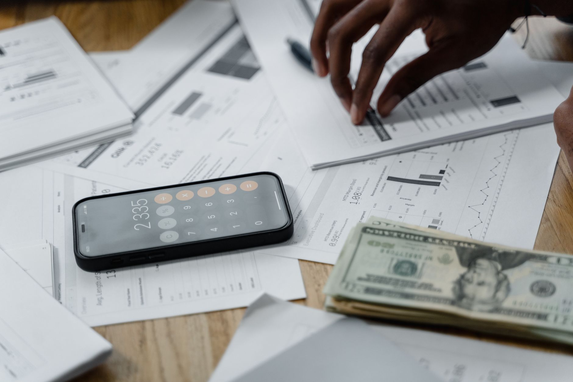 Close-up of hands calculating financial data with reports, smartphone, and cash on table.