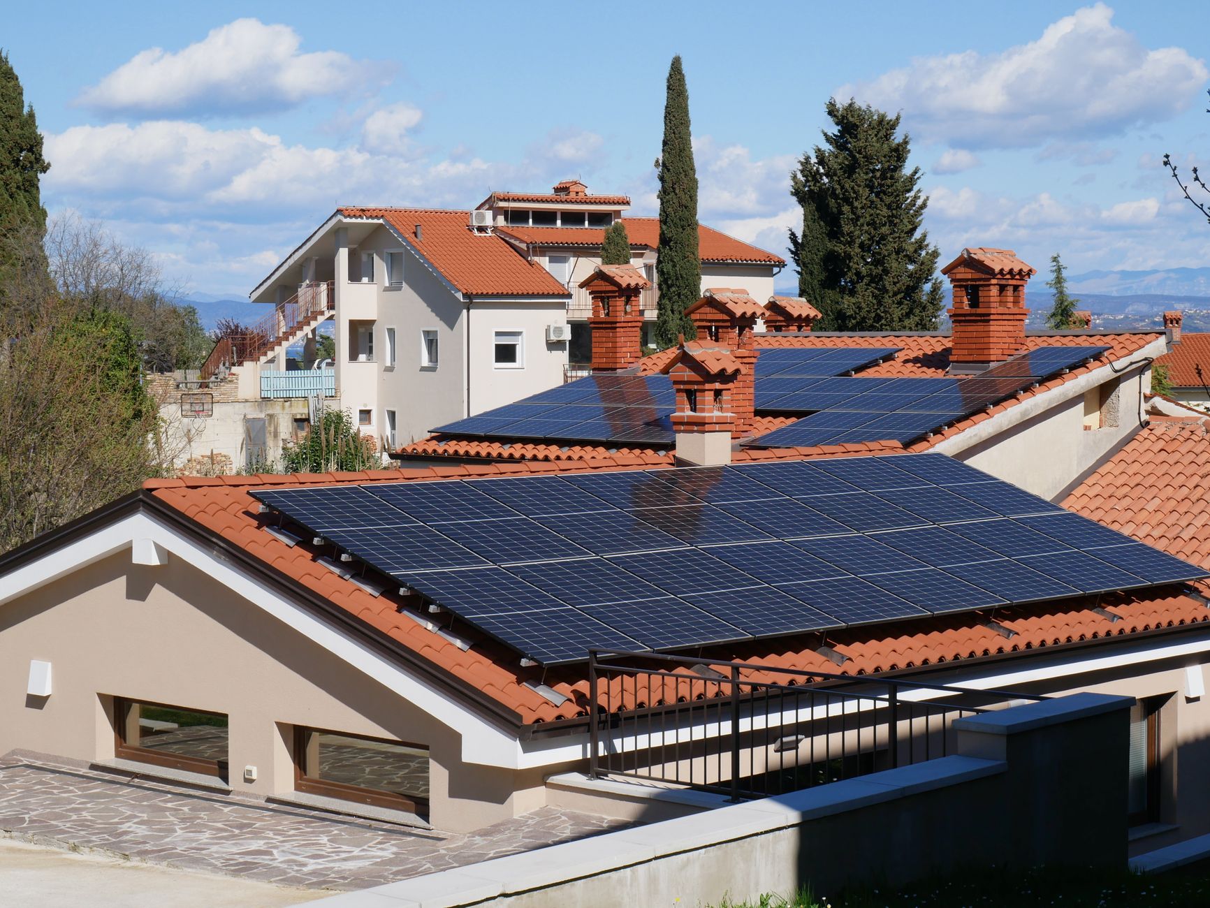 A panoramic view of solar panels on red tile rooftops in a sunny suburban neighborhood.