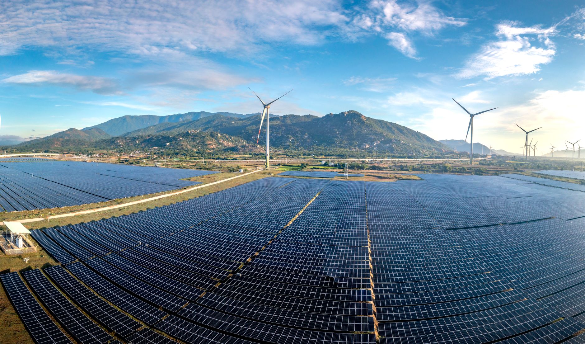 Aerial view of a vast solar farm with wind turbines against a mountainous backdrop on a sunny day.