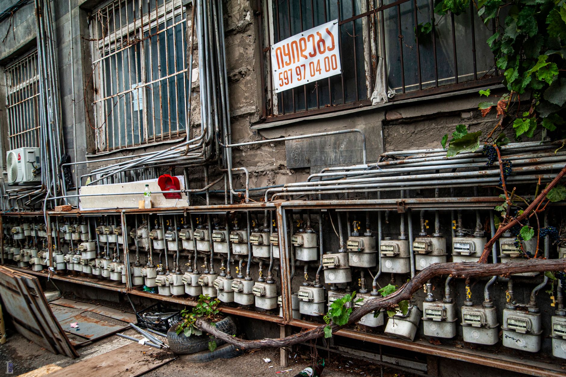 Row of outdoor gas meters on a building facade with vines and a sign in non-Latin script.