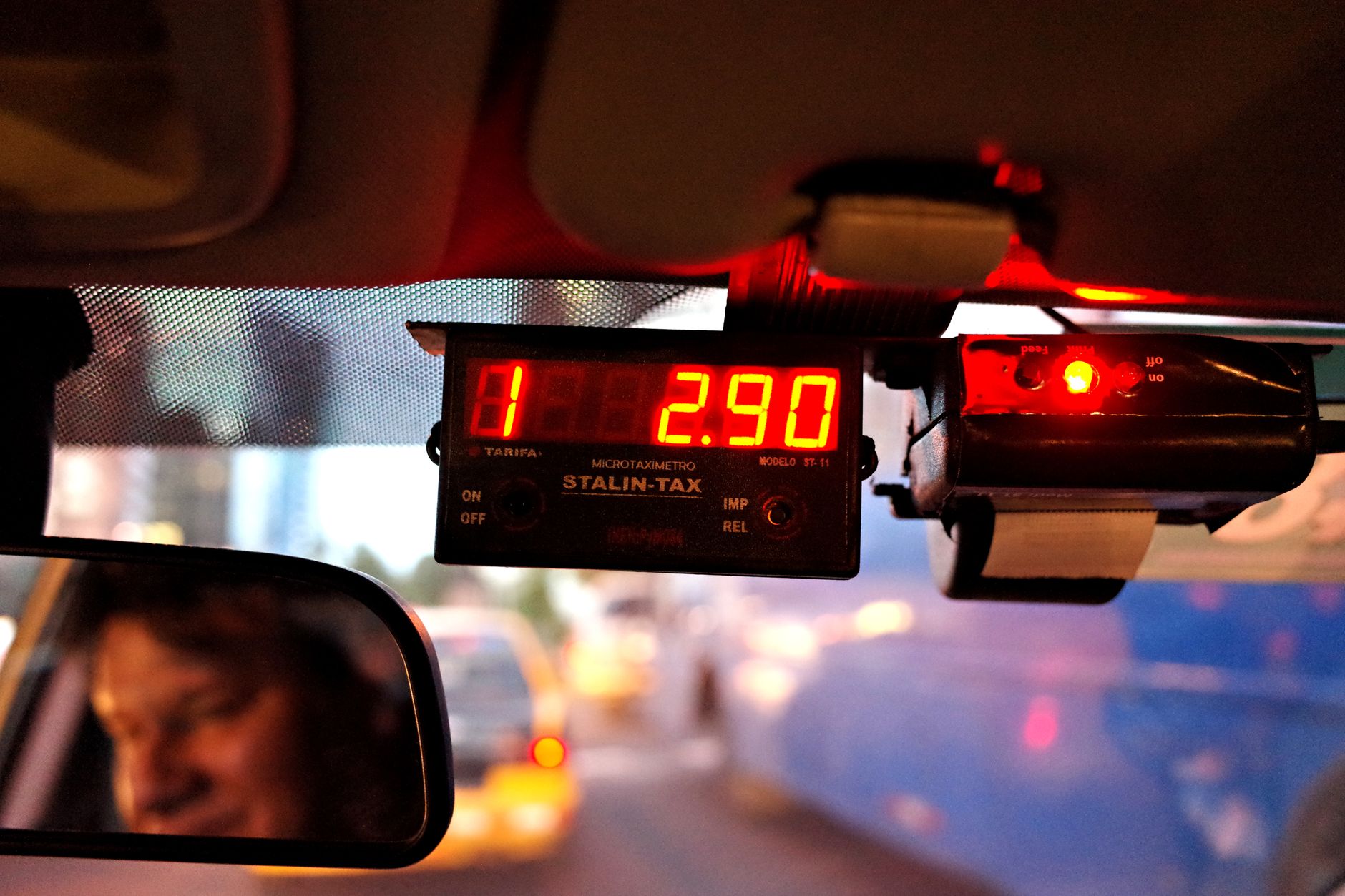 Taxi meter showing fare in city traffic with driver's reflection visible.