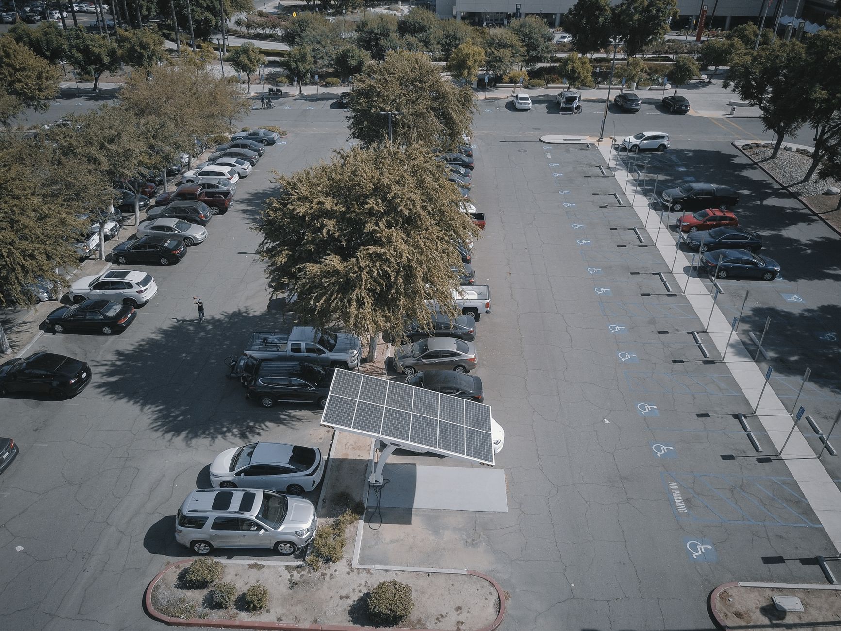 Aerial view of a solar panel charging station in a busy parking lot surrounded by cars and trees.
