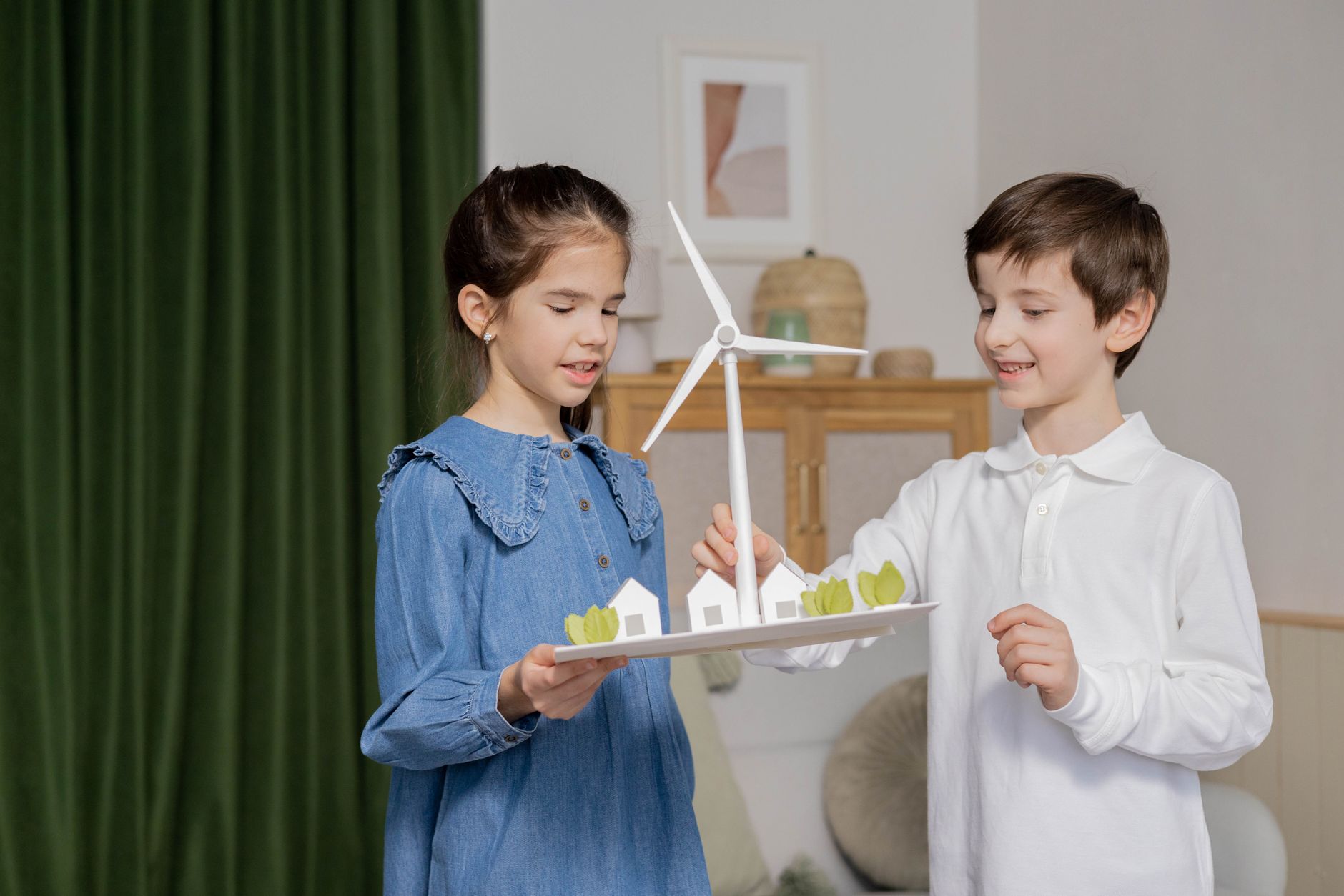 Two children holding a wind turbine diorama indoors, emphasizing renewable energy education.