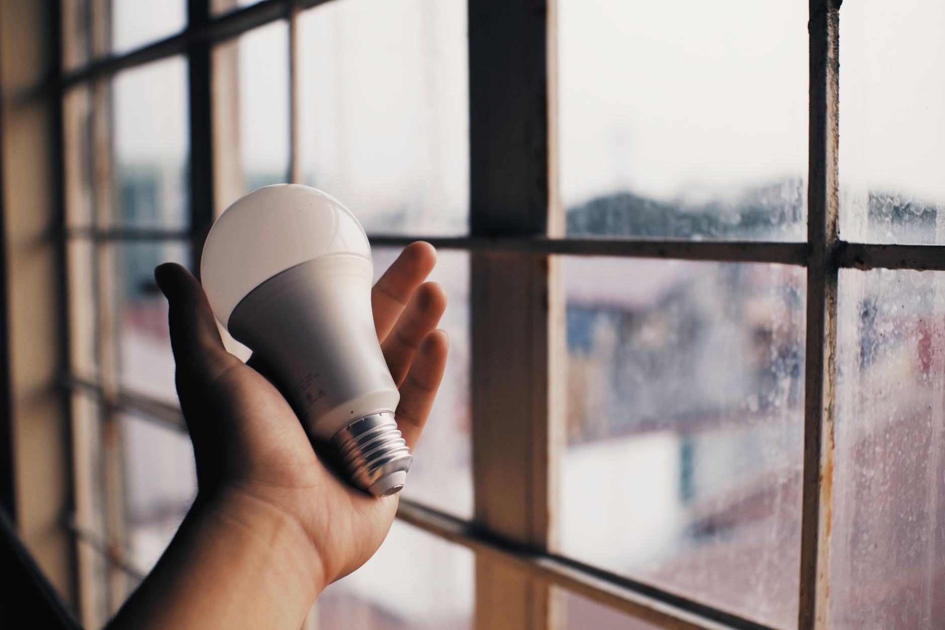 Anonymous person holding energy efficient light bulb in hand on blurred background of big dusty window in light room